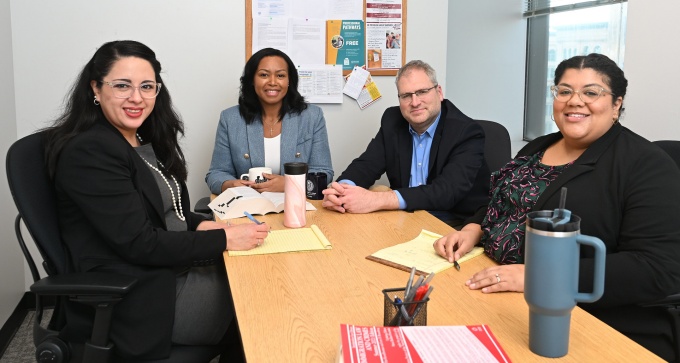 Four people sit around a rectangular wooden table in an office meeting room. The table has yellow notepads, pens, and reusable drink containers. A bulletin board with various notices and flyers is mounted on the wall behind them, and a window with blinds is on the right side of the room.