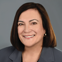 Person with straight, shoulder-length dark brown hair wearing a dark gray blazer over a black top, accessorized with small hoop earrings and a thin necklace, posed against a plain gray background.