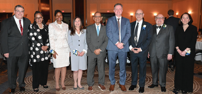 Group of nine people standing in a line at a formal indoor event. They are dressed in business or evening attire, including suits, ties, and dresses. Several individuals wear corsages or boutonnieres with blue and white flowers. The background shows banquet tables and warm-toned walls with decorative panels. Seven people posed indoors around a round dining table set with glasses of water, iced tea, and bread rolls. Four individuals are standing behind the table wearing suits and collared shirts, and three are seated in front wearing sweaters or jackets. The background features colorful abstract wall art with circular designs and metallic accents.