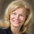 Person with shoulder-length blonde hair wearing a black jacket over a light blue top and a chunky necklace featuring large purple and light green beads. The background is a smooth gradient in neutral tones.