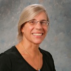 Person with gray hair pulled back, wearing a black V-neck shirt and a gold necklace with a blue pendant. The background is a textured gray backdrop with subtle blue and beige tones.