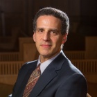 Person wearing a dark navy suit jacket, white dress shirt, and a patterned tie with red and gold tones, seated on a wooden bench in a softly lit interior space with wooden architectural details in the background.