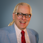 Person wearing a light blue suit jacket over a pale pink dress shirt with a red tie featuring a blue dot pattern. The background is a smooth gradient in shades of blue.