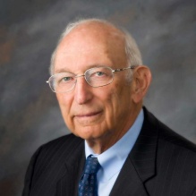Person wearing a dark pinstripe suit with a light blue dress shirt and a patterned blue tie. A small American flag lapel pin is visible on the left side of the suit jacket. The background is a softly blended gray backdrop.