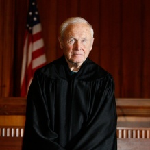 Person wearing a black judicial robe standing in a courtroom. Behind them is a wooden judge’s bench with an American flag and a lit lamp visible in the background. The setting includes wood-paneled walls and a formal courtroom atmosphere.