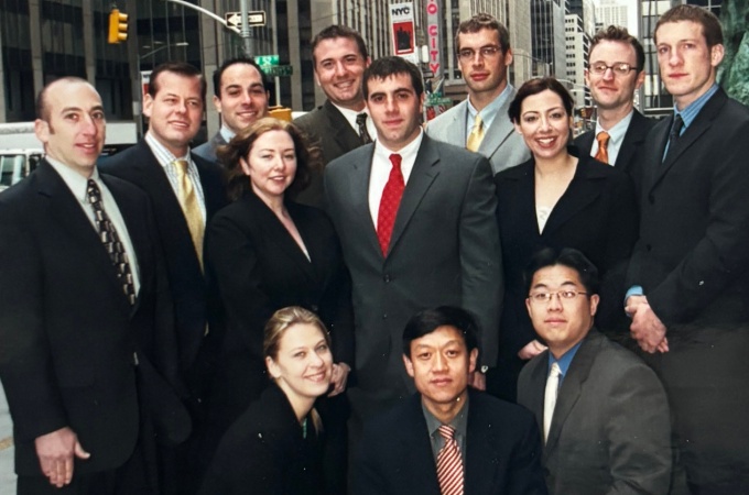 A group of people dressed in formal business attire standing together on a city sidewalk in an urban setting. Tall office buildings line the street, and the Radio City Music Hall sign is visible in the background. The group appears posed for a photo, with some standing and others kneeling in front.