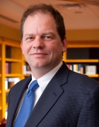 Person wearing a dark checkered suit jacket, white dress shirt, and a textured blue tie, standing in front of yellow bookshelves filled with books in an indoor setting with warm lighting.