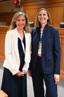Two people standing in a courtroom setting with wooden judge’s bench and chairs in the background. One person is wearing a white blazer over a dark dress, and the other is wearing a navy pantsuit with a patterned blouse.