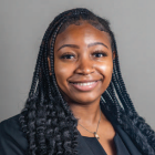 Studio portrait of a person with long braided hair, wearing a dark blazer and a necklace, photographed against a muted green background.