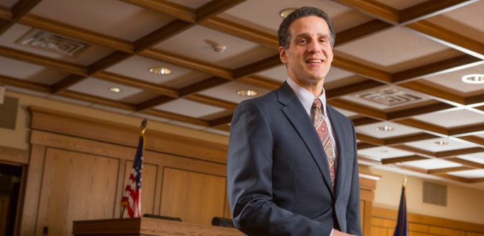 man wearing a suit, sitting on a bench in a courtroom.