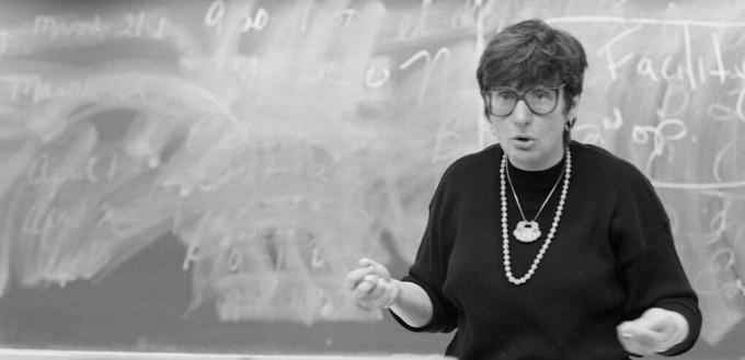 black and white photo of woman wearing black sweater in front of a chalkboard.