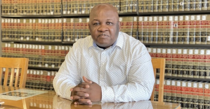 black gentleman wearing white shirt, sitting at a wooden table with shelved books behind him.