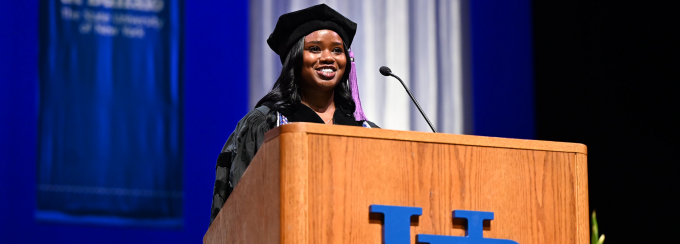 young woman wearing commencement regalia, standing at a podium.
