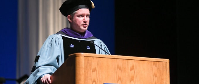 Todd Brown, dressed in graduation regalia, speaking at a podium.