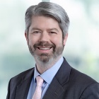 man smiling, standing in front of a large window.