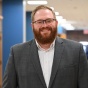 man wearing glasses, smiling, standing inside a school hallway. 