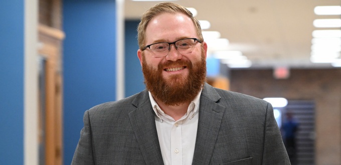 man wearing glasses, smiling, standing inside a school hallway. 