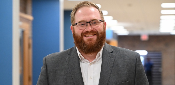man wearing glasses, smiling, standing inside a school hallway.