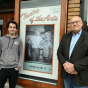 Two men, smiling, standing outside next to a movie poster. 