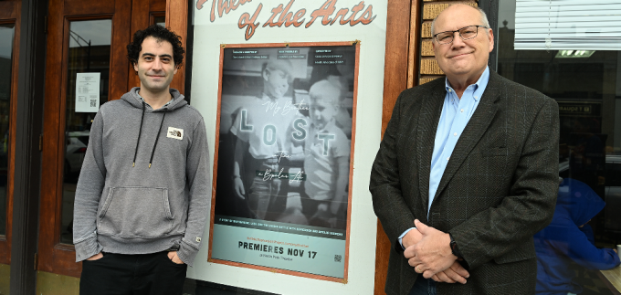 Two men, smiling, standing outside next to a movie poster. 