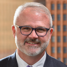 a man wearing glasses, smiling, standing in front of a window.