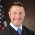 man standing in front of US flag, wearing a judges robe, smiling.