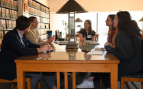 A group of students in a library, sitting around a table, talking. 