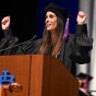 Young woman wearing graduation regalia, standing at a podium, waving. 