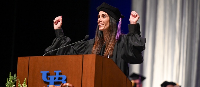 Young woman wearing graduation regalia, standing at a podium, waving. 