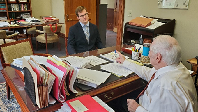 Man sitting at a cluttered desk acrossed from a youn man in a suit. 