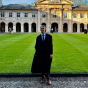 Man wearing a black robe, posing outside, in a grassy university quad. 