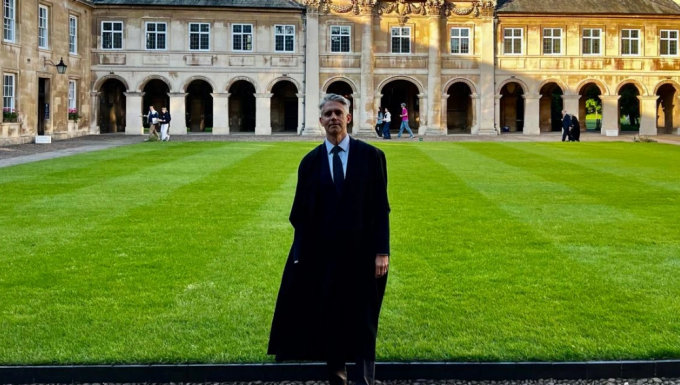 Man wearing a black robe, posing outside, in a grassy university quad. 