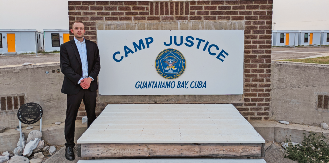 man standing outside next to a sign that says Camp Justice Guantanamo Bay, Cuba. 