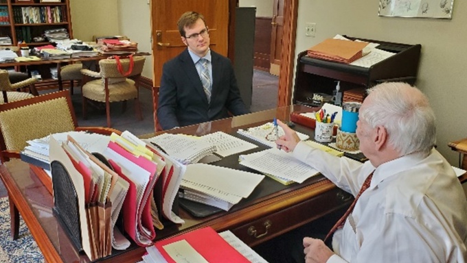 Man sitting at a cluttered desk talking to a young man sitting opposite. 