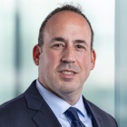 man wearing a suit and dark colored tie, standing in front of a large window.