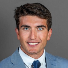 Young man wearing light blue colored suit, dark blue tie, smiling.
