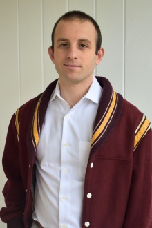 Young man wearing burgundy wool jacket, white button-down shirt, standing in a hallway.