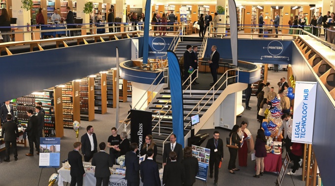 A large atrium for a library, filled with people standing around tables, talking to one another. 