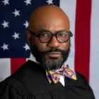 A man in judges robes, wearing yellow and puple striped bow tie, standing in front of a US flag. 