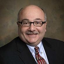 Man wearing dark blazer, tie, glasses, smiling. 