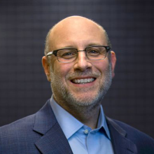 Man wearing blue blazer, blue shirt, and glasses, smiling. 