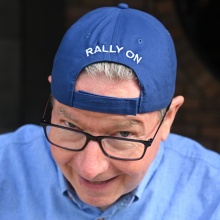 Closeup image of Mac Donald wearing backwards baseball cap that includes text that says Rally On.