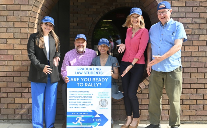 A small group of people wearing blue baseball caps, standing outside holding a poster for Rally the Pass.
