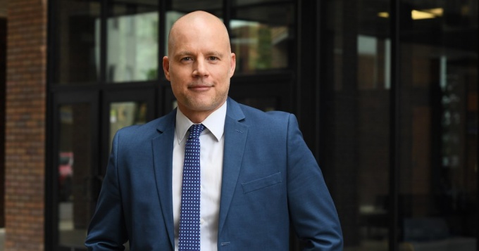 Man standing outside a building, wearing suit and tie, smiling.