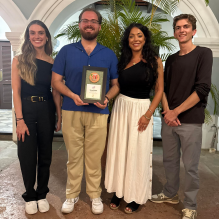 Four people standing together indoors in front of green plants and an arched doorway. One person in the center is holding a rectangular award plaque with a circular emblem and text. They are dressed in semi-formal attire, including black, white, blue, and gray outfits.