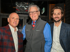 Three people standing together indoors at an event, wearing semi-formal attire including blazers and a vest, with a dark background and glass doors behind them.