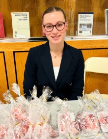 A person is seated behind a table displaying multiple clear plastic bags filled with red and white striped candy canes. In front of the table is a sign with the text “Leaders for Philanthropy” and a crest logo. Behind the person is a wooden display case with shelves holding various law-related books and journals.