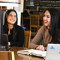 Two people sit at a table in a library or study area with open laptops and a book between them. They appear to be engaged in conversation. Bookshelves and wooden chairs are visible in the background, suggesting an academic setting.
