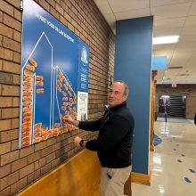 Person standing in a hallway next to a brick wall, placing an orange sticker on a large blue poster titled “Share Your UB Law Blueprint.” The poster features an outline of a building with many orange stickers already attached, and includes text about the UB Law Blueprint initiative. The hallway has beige floors, blue columns, and overhead fluorescent lighting.