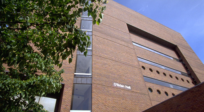 Exterior view of a multi-story brick building with narrow horizontal windows and circular window details. A vertical glass section runs along the left side, partially framed by leafy tree branches in the foreground. The building name “O’Brian Hall” is visible on the brick facade under a clear blue sky.